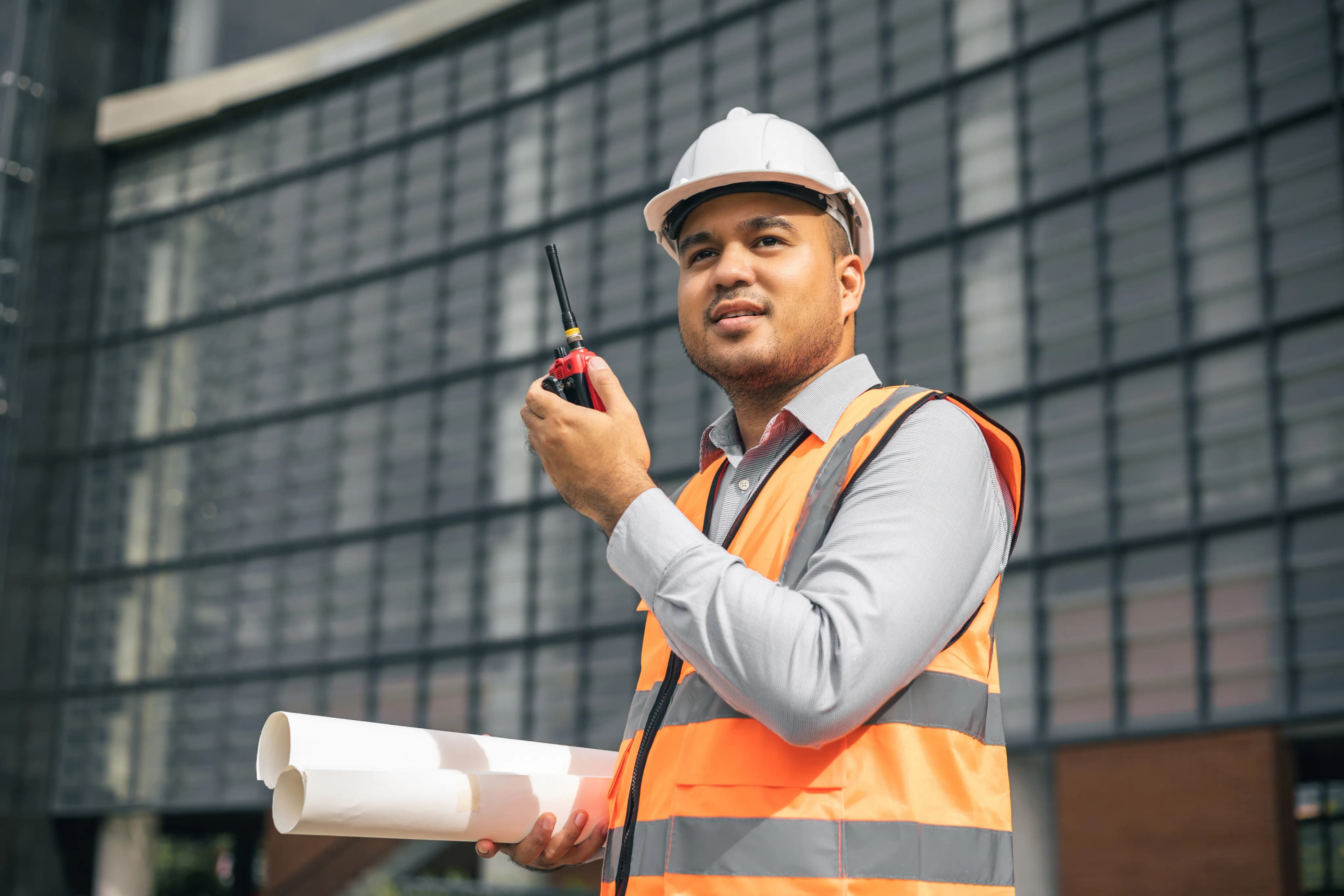 Site engineer in safety gear holding blueprints and a radio outside a modern building
